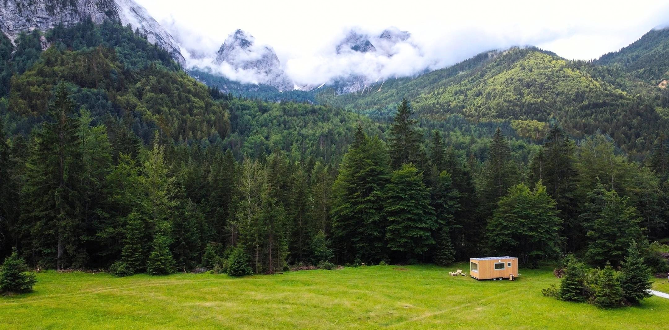 Casetta Friland in legno in una radura tra gli abeti, con le montagne sullo sfondo